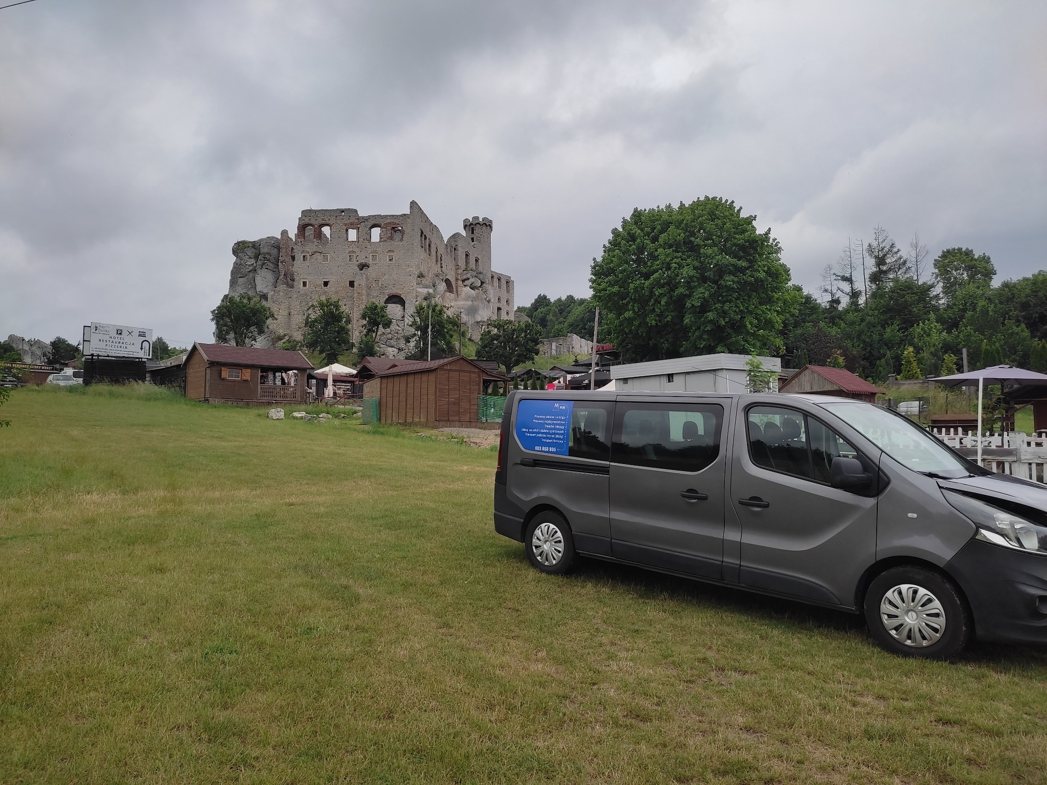 Szary bus transportowy z reklamą na boku zaparkowany na trawiastym terenie, w tle ruiny zamku Ogrodzieniec pod pochmurnym niebem.