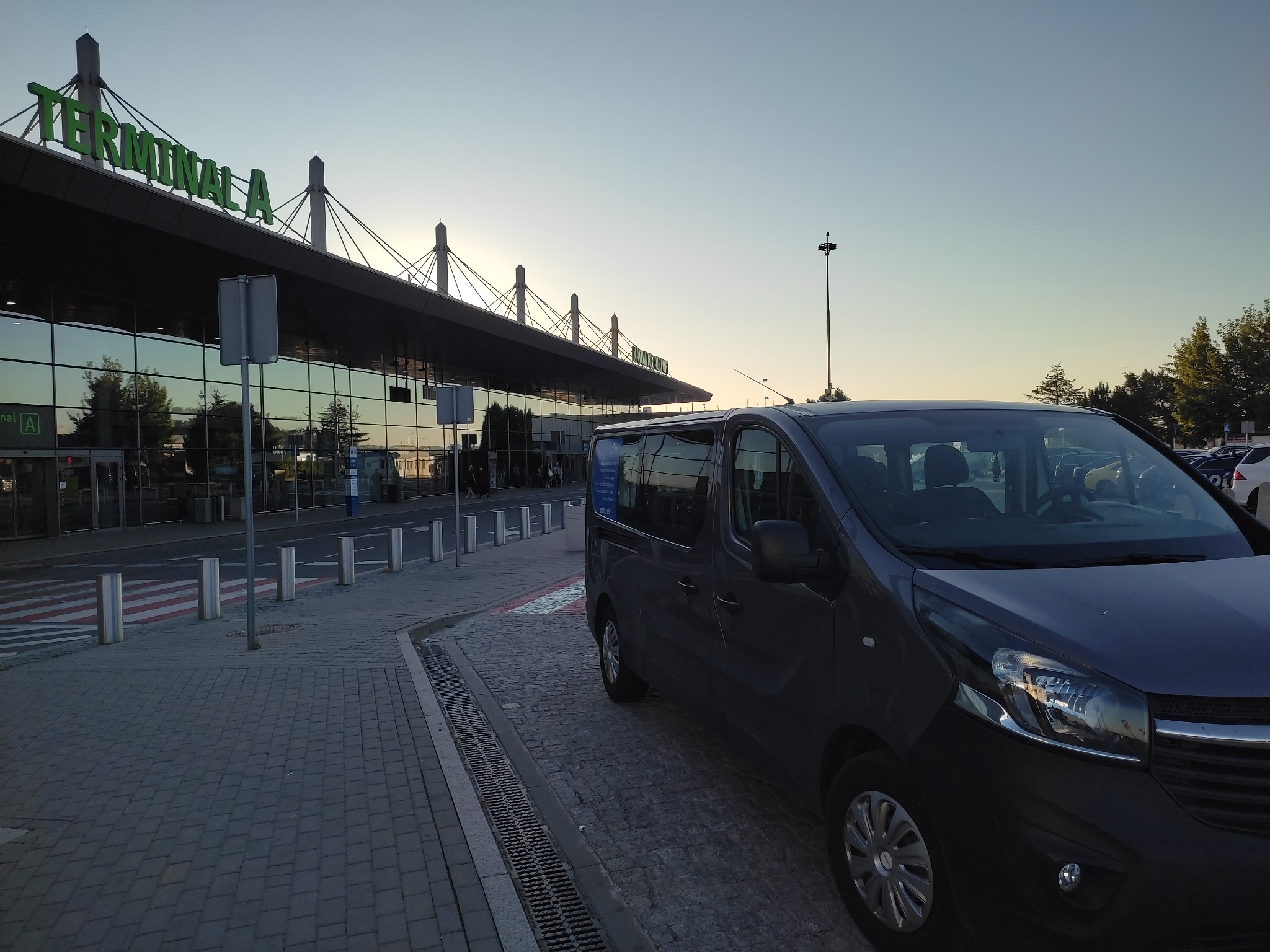 Szary van zaparkowany przed terminalem lotniska o wschodzie słońca, widoczny napis Terminal A na budynku.