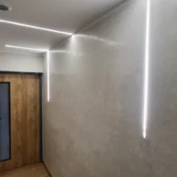 Wykończony korytarz z oświetleniem LED wzdłuż sufitu i pionowo na ścianie, z teksturą na ścianie i drewnianymi drzwiami z szybą. Nowoczesny design.