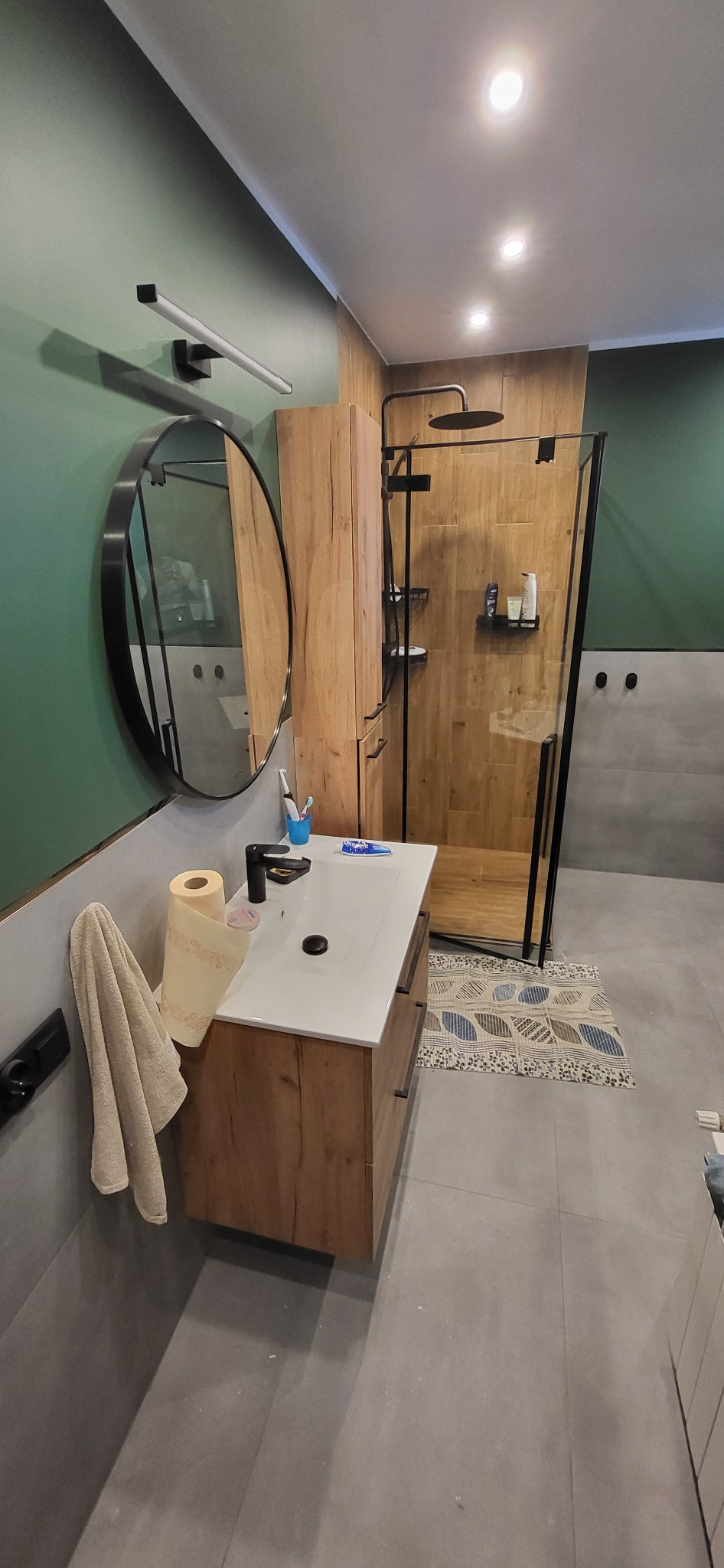 Nowoczesna łazienka z kabiną prysznicową walk-in, drewnianą szafką pod umywalką i okrągłym lustrem w Proszowicach. Zielona ściana i szara podłoga.