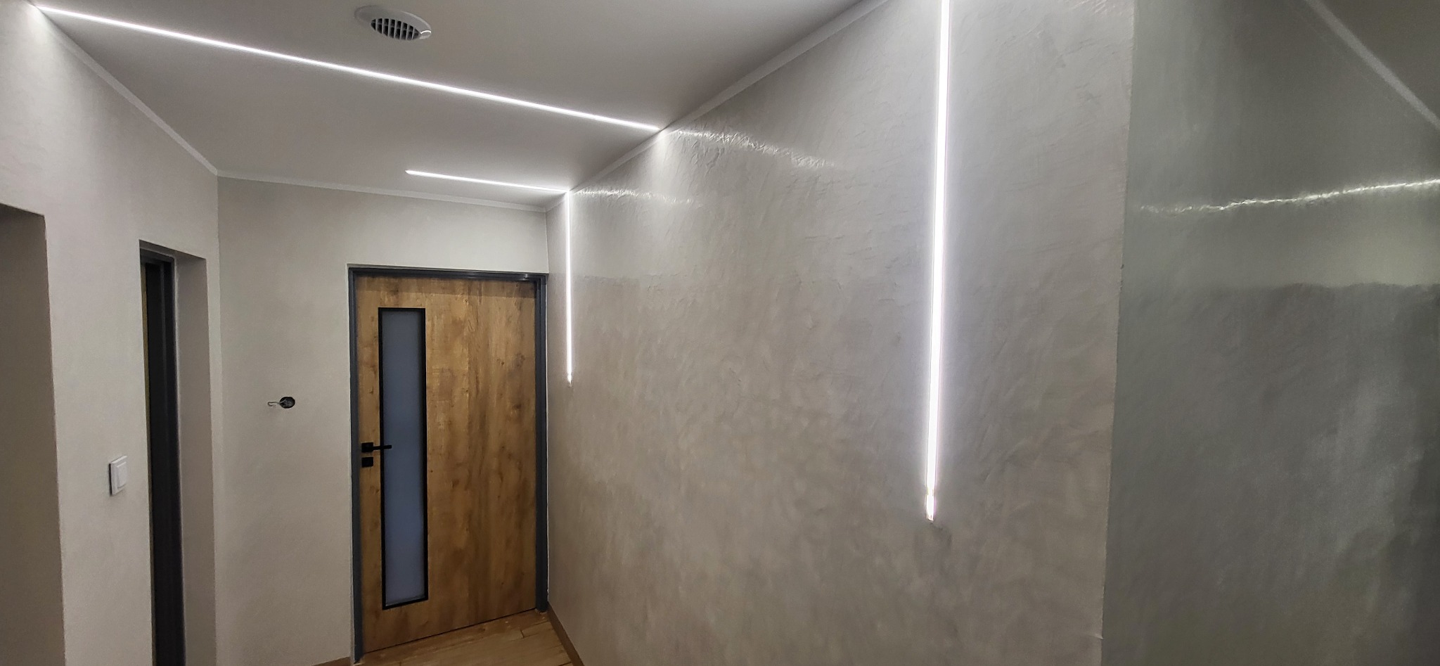 Wykończony korytarz z oświetleniem LED wzdłuż sufitu i pionowo na ścianie, z teksturą na ścianie i drewnianymi drzwiami z szybą. Nowoczesny design.