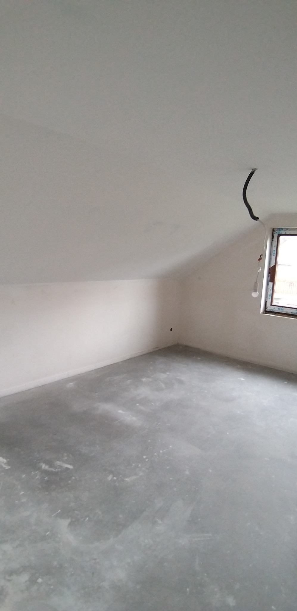 Poddasze po szpachlowaniu, widoczne gładkie ściany i przygotowana podłoga. Okno z PCV w prawym rogu, przewody elektryczne zwisają z sufitu.