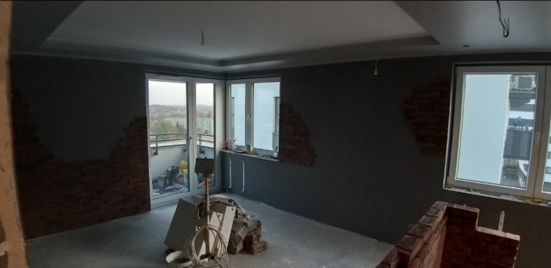Wnętrze w trakcie wykańczania: szare ściany z fragmentami cegieł dekoracyjnych, okna i balkon. Widoczne narzędzia i materiały budowlane.