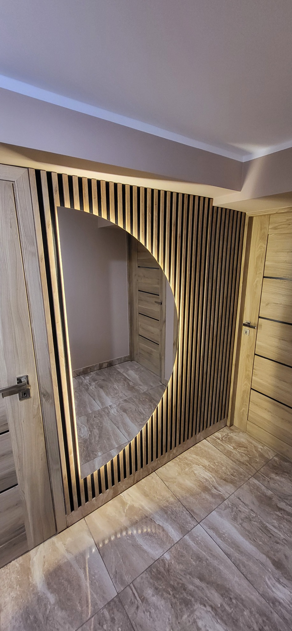 Nowoczesna aranżacja wnętrza z drewnianą ścianką działową z półokrągłym otworem, podświetloną ciepłym światłem. Widoczna podłoga z płytek imitujących marmur.