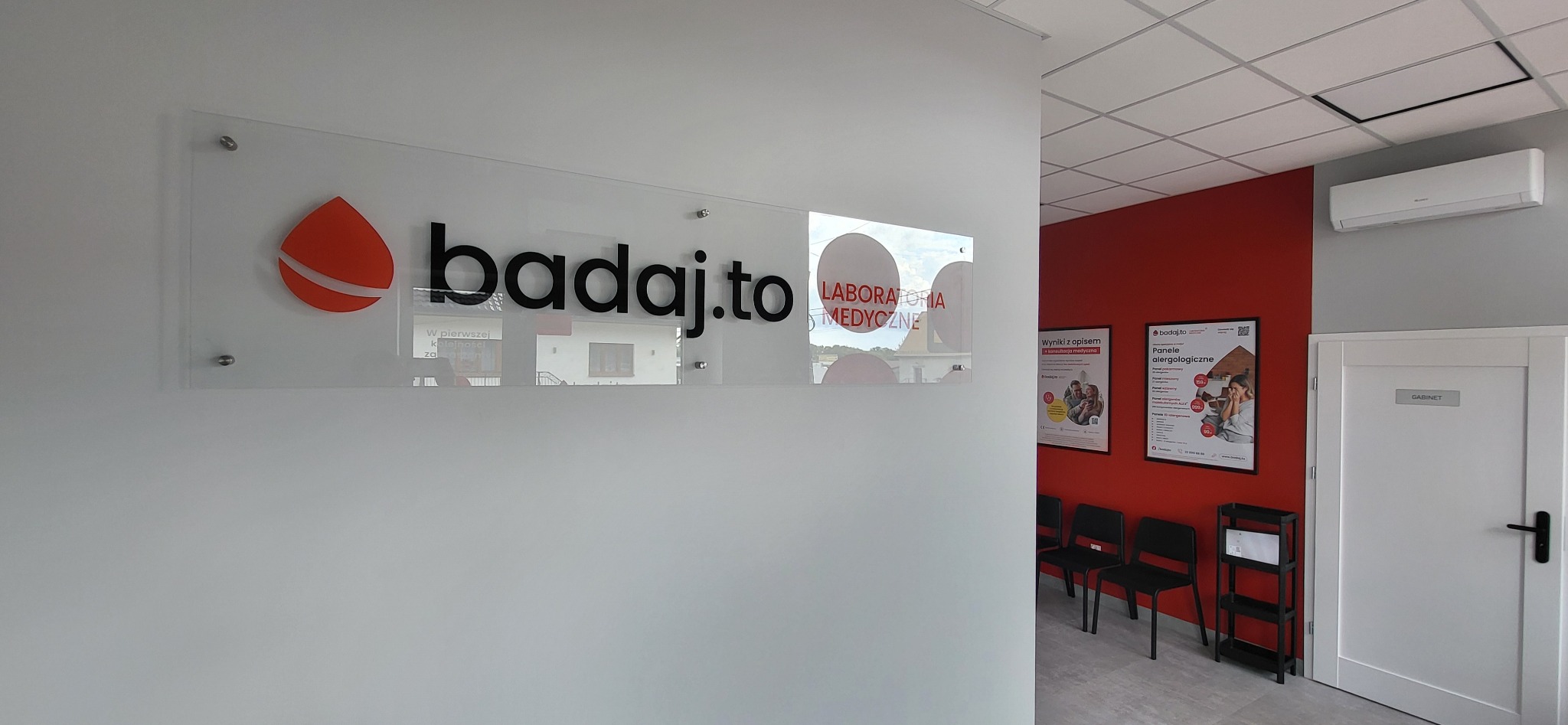Nowoczesne oznakowanie: logo 'badaj.to' na przezroczystej płycie, z widocznym wnętrzem laboratorium medycznego w tle, minimalistyczny design i oświetlenie.