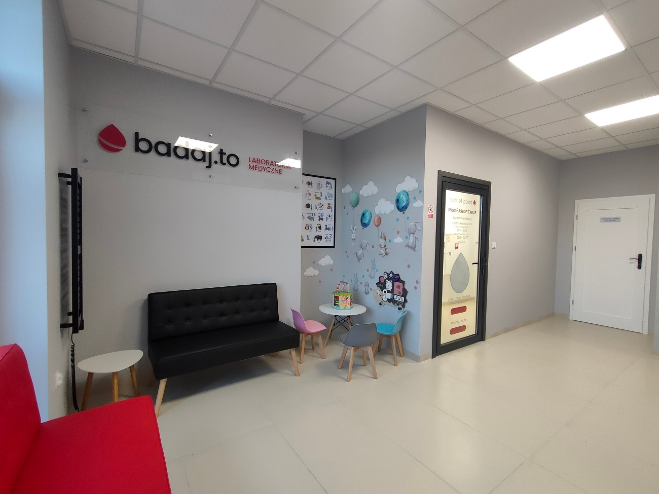 Jasne, nowoczesne wnętrze poczekalni laboratorium medycznego z czarną sofą, stolikami i krzesłami dla dzieci, dekorowane naklejkami na ścianie.