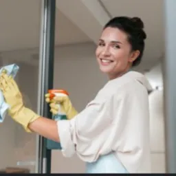 Uśmiechnięta kobieta w żółtych rękawicach, z błękitną ściereczką i spryskiwaczem, czyści szklane drzwi w jasnym wnętrzu.