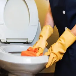 Osoba w uniformie i żółtych rękawicach czyści muszlę klozetową pomarańczową ściereczką, widoczny kosz wiklinowy.