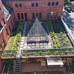 Astall sp. z o.o. - Widok z góry na zielony dach z piramidalną szklaną konstrukcją, otoczony ceglanymi budynkami, z metalową balustradą i schodami prowadzącymi na niższy poziom.