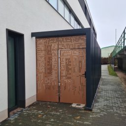 Astall sp. z o.o. - Nowoczesna, ażurowa brama wjazdowa z metalu w kolorze miedzianym, z regularnym wzorem perforacji, zamontowana przy budynku z jasną elewacją i ciemnymi ramami okiennymi, widoczna z zewnątrz...