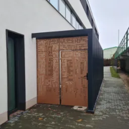 Nowoczesna, ażurowa brama wjazdowa z metalu w kolorze miedzianym, z regularnym wzorem perforacji, zamontowana przy budynku z jasną elewacją i ciemnymi ramami okiennymi, widoczna z zewnątrz...