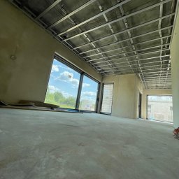 REMONT-BUD Jakub Ćwikłowski - Wnętrze w stanie deweloperskim z widoczną konstrukcją sufitu podwieszanego z profili metalowych, surowe ściany przygotowane do dalszych prac wykończeniowych, duże okna z widokiem na zieleń...