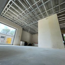 REMONT-BUD Jakub Ćwikłowski - Wnętrze w trakcie prac wykończeniowych: widoczna metalowa konstrukcja podwieszanego sufitu, ściany po wstępnym szpachlowaniu, duże okno z widokiem na zewnątrz. Na podłodze butelka wody mineralnej.
