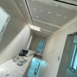 REMONT-BUD Jakub Ćwikłowski - Korytarz w trakcie remontu: ściany i sufit pokryte warstwą szpachli, widoczne łączenia płyt karton-gips, puszka z masą szpachlową Knauf Super Finish na podłodze, czarny worek i resztki materiałów...