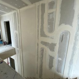 REMONT-BUD Jakub Ćwikłowski - Wnętrze w trakcie remontu: ściany z płyt kartonowo-gipsowych z dekoracyjnymi elementami w kształcie łuków, częściowo pokryte masą szpachlową, widoczne schody i otwór drzwiowy.