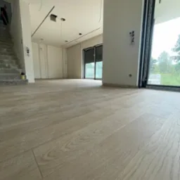 Jasna, drewniana podłoga ułożona w przestronnym, surowym wnętrzu z widocznymi przewodami elektrycznymi wystającymi z sufitu, schodami w stanie surowym i dużym oknem z widokiem na zieleń.