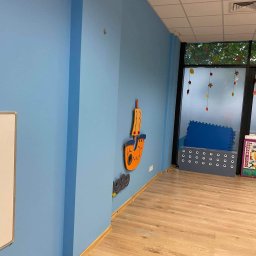 REMONT-BUD Jakub Ćwikłowski - Świeżo pomalowane na błękitno ściany w pokoju dziecięcym z drewnianą podłogą, ozdobione drewnianymi dekoracjami ściennymi w kształcie łodzi i fal. W tle widoczne okno z jesiennymi dekoracjami...