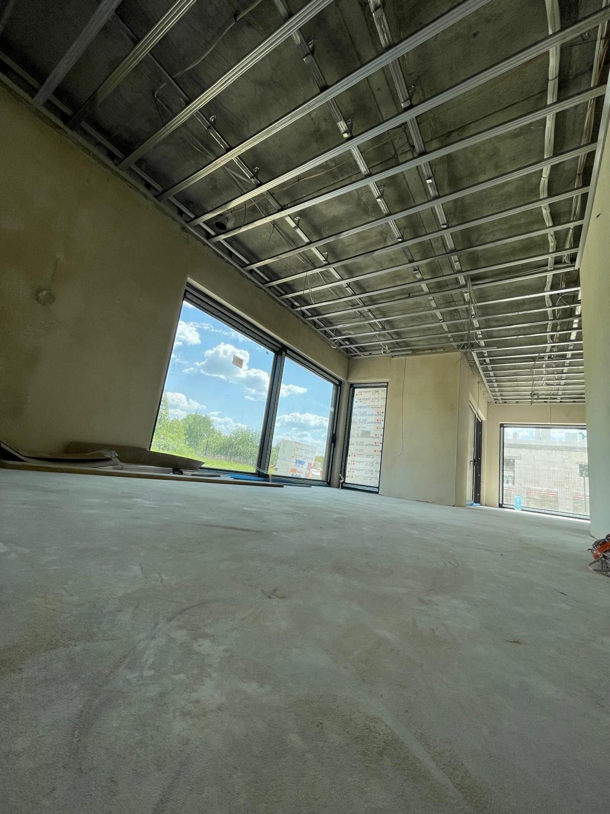 Wnętrze w stanie deweloperskim z widoczną konstrukcją sufitu podwieszanego z profili metalowych, surowe ściany przygotowane do dalszych prac wykończeniowych, duże okna z widokiem na zieleń...