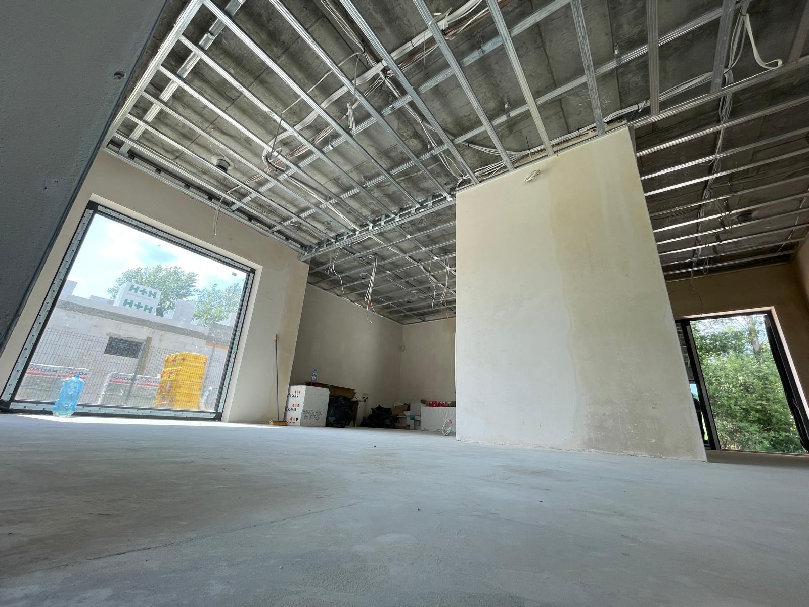 Wnętrze w trakcie prac wykończeniowych: widoczna metalowa konstrukcja podwieszanego sufitu, ściany po wstępnym szpachlowaniu, duże okno z widokiem na zewnątrz. Na podłodze butelka wody mineralnej.