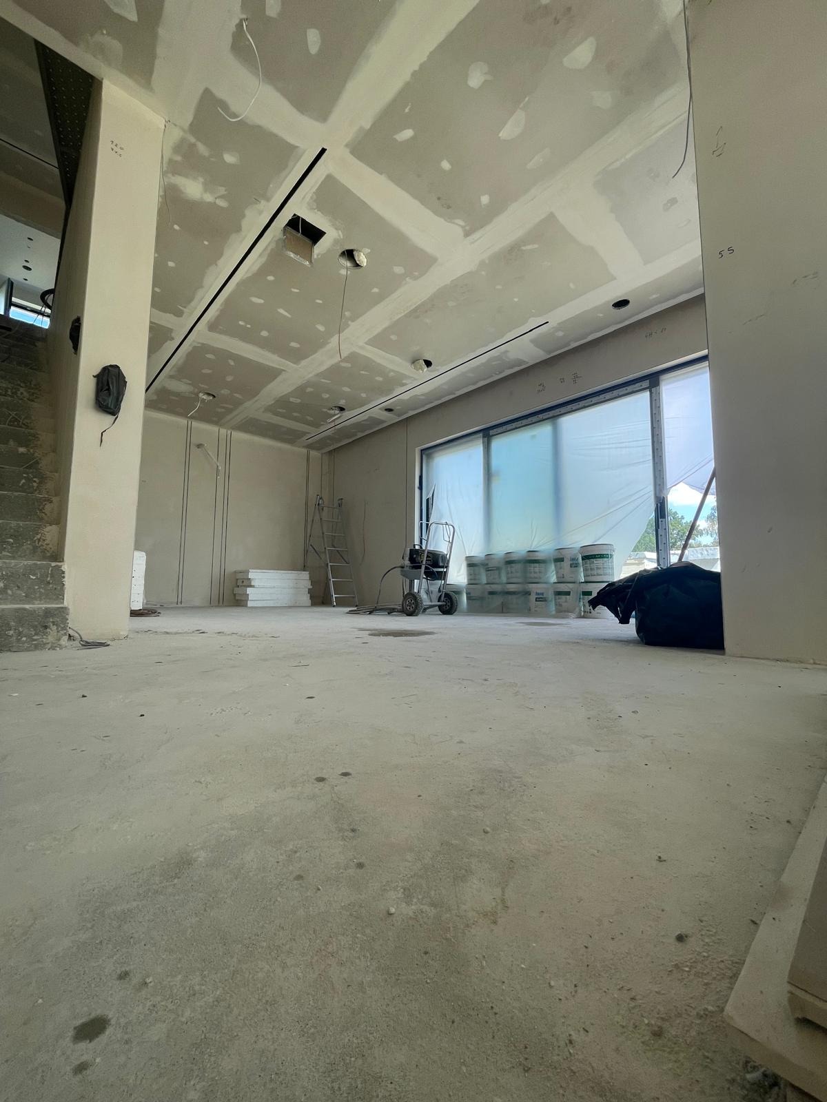 Wnętrze w trakcie prac wykończeniowych: sufit z płyt kartonowo-gipsowych z widocznymi łączeniami, ściany przygotowane do dalszej obróbki, podłoga surowa, widoczne narzędzia i materiały budowlane...