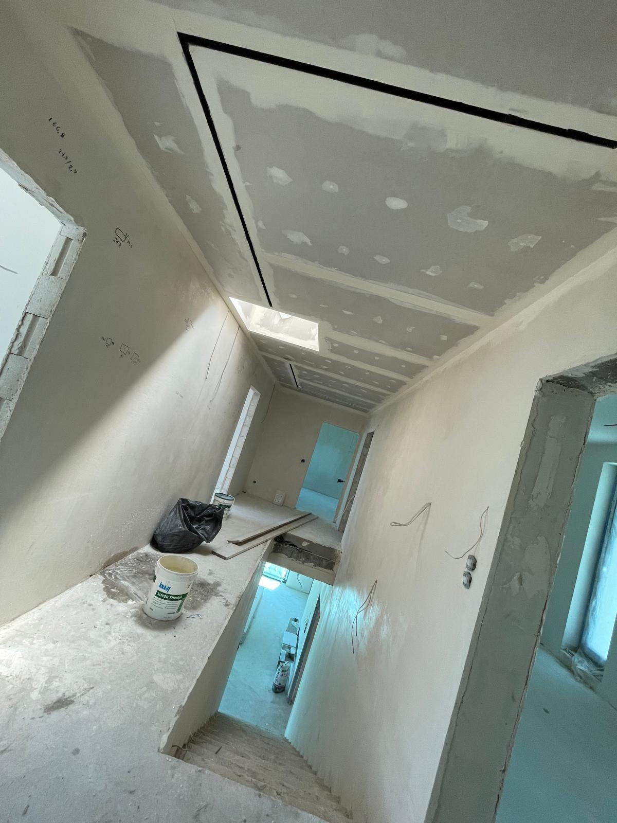 Korytarz w trakcie remontu: ściany i sufit pokryte warstwą szpachli, widoczne łączenia płyt karton-gips, puszka z masą szpachlową Knauf Super Finish na podłodze, czarny worek i resztki materiałów...