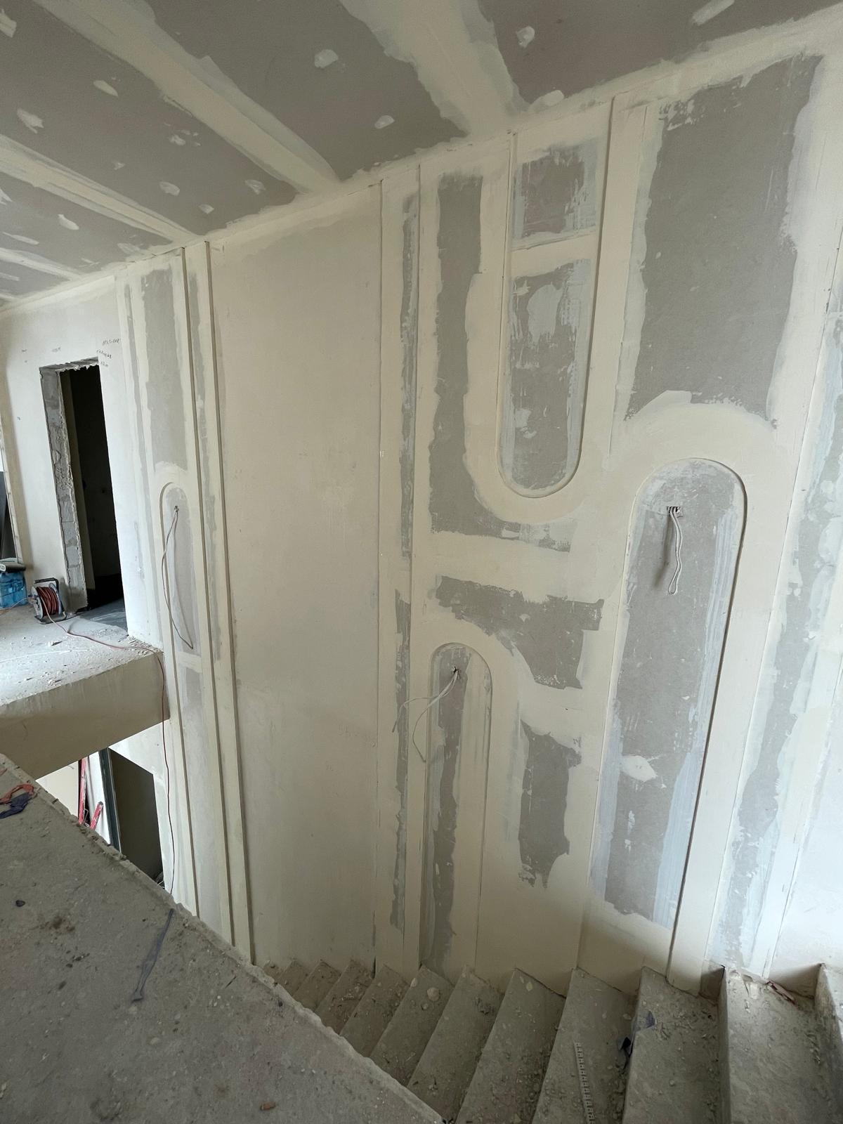 Wnętrze w trakcie remontu: ściany z płyt kartonowo-gipsowych z dekoracyjnymi elementami w kształcie łuków, częściowo pokryte masą szpachlową, widoczne schody i otwór drzwiowy.