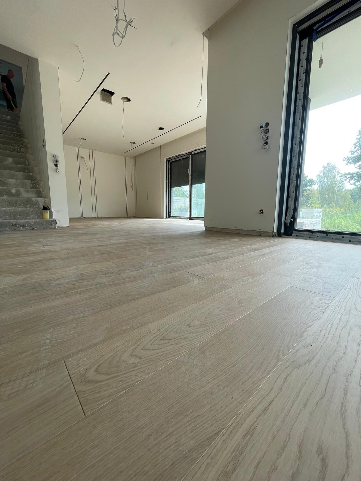 Jasna, drewniana podłoga ułożona w przestronnym, surowym wnętrzu z widocznymi przewodami elektrycznymi wystającymi z sufitu, schodami w stanie surowym i dużym oknem z widokiem na zieleń.