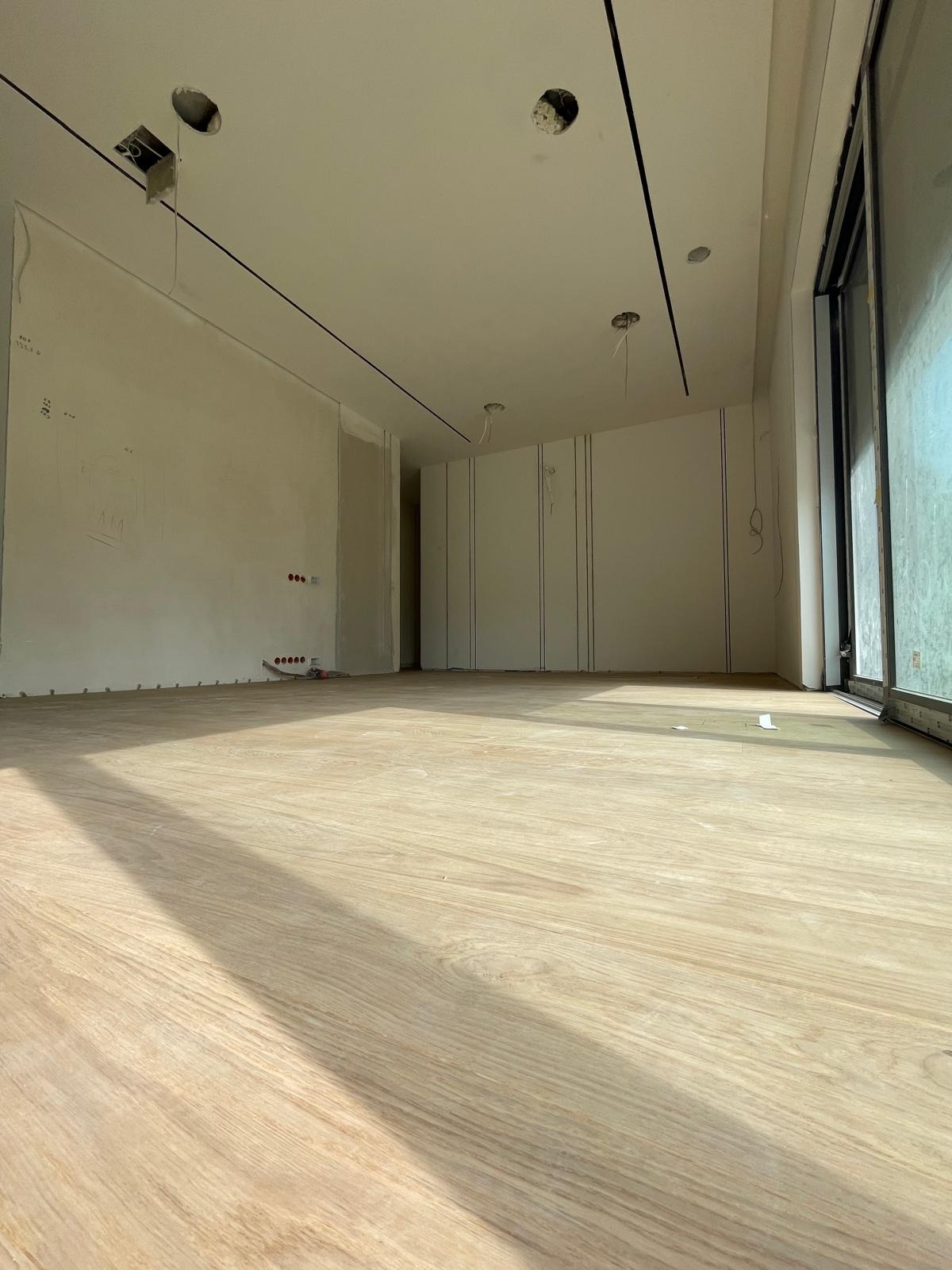 Wnętrze w stanie deweloperskim z drewnianą podłogą, przygotowane do prac wykończeniowych, widoczne niedokończone otwory w suficie na oświetlenie, pionowe rowki na ścianie i instalacja elektryczna.