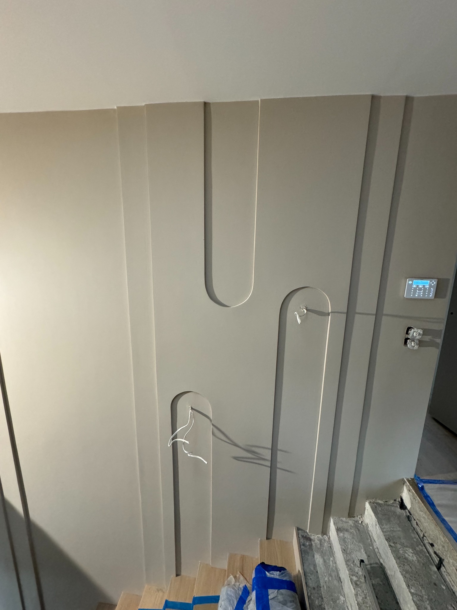 Ściana z ozdobnymi panelami w odcieniach szarości, z widocznym okablowaniem, elementami instalacji elektrycznej i zabezpieczonymi schodami, w trakcie prac wykończeniowych.