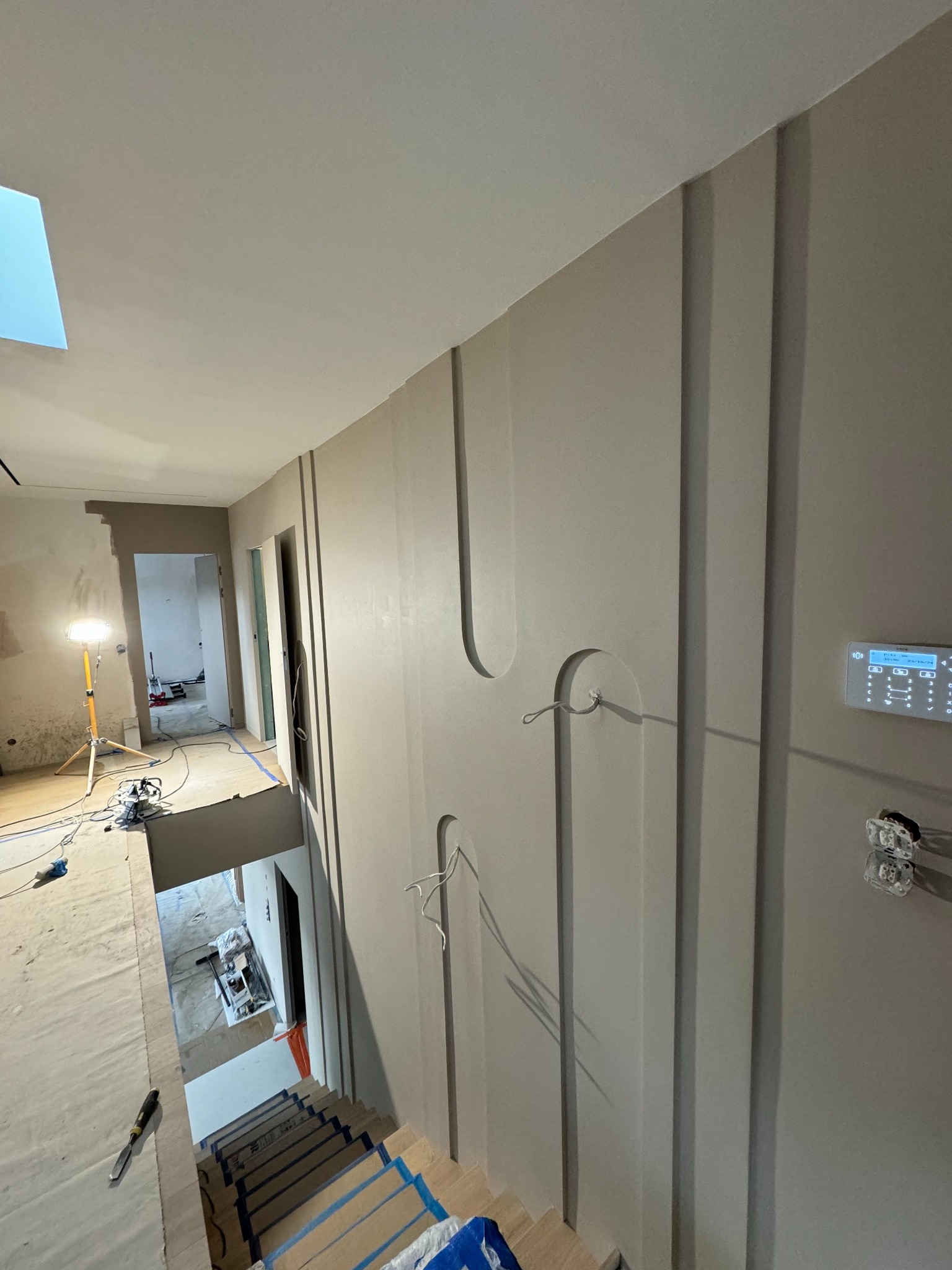 Wnętrze w trakcie wykańczania: świeżo pomalowana ściana z dekoracyjnymi pionowymi panelami i półokrągłymi wnękami, widoczne schody zabezpieczone folią, narzędzia na podłodze i lampa oświetlająca...