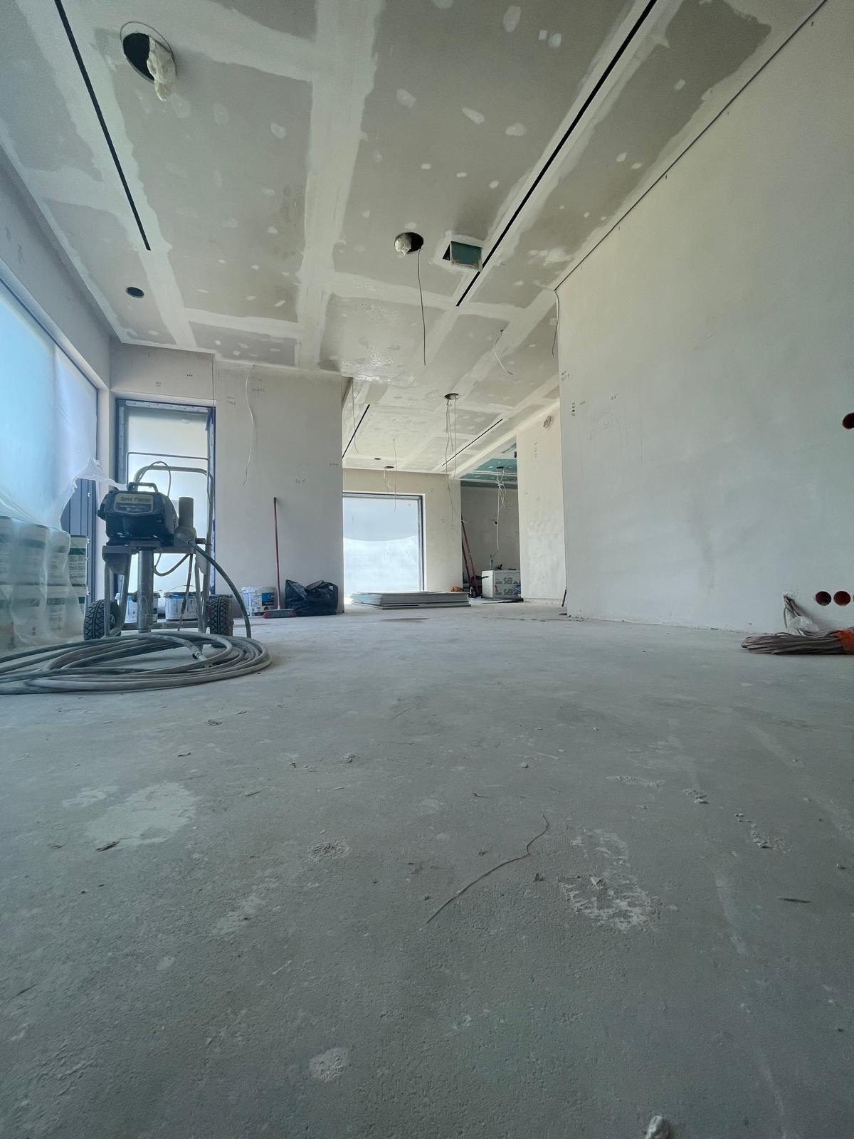 Wnętrze w trakcie prac wykończeniowych: ściany przygotowane, częściowo wyszpachlowane, widoczna maszyna do malowania natryskowego, sufity podwieszane z płyt gipsowo-kartonowych z widocznymi...
