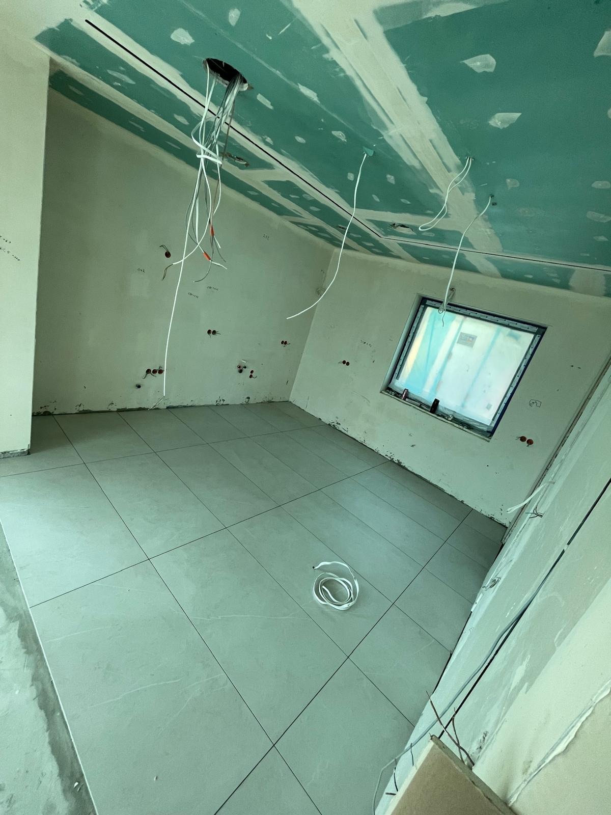 Pokój w trakcie remontu: sufit i ściany pokryte płytami gipsowo-kartonowymi, widoczne kable wystające z sufitu, świeżo położona jasna podłoga z płytek, okno z widokiem na zewnątrz.