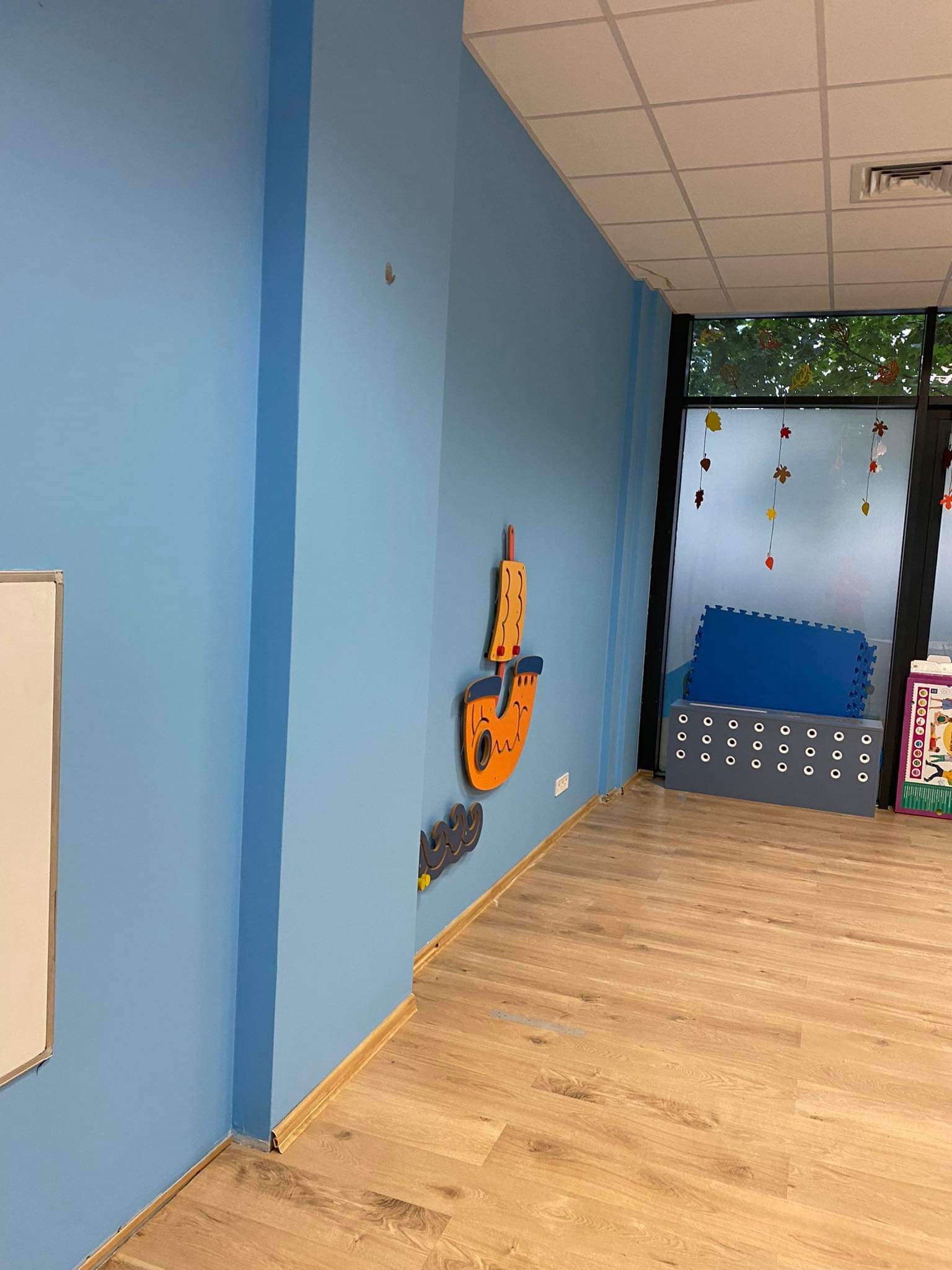 Świeżo pomalowane na błękitno ściany w pokoju dziecięcym z drewnianą podłogą, ozdobione drewnianymi dekoracjami ściennymi w kształcie łodzi i fal. W tle widoczne okno z jesiennymi dekoracjami...