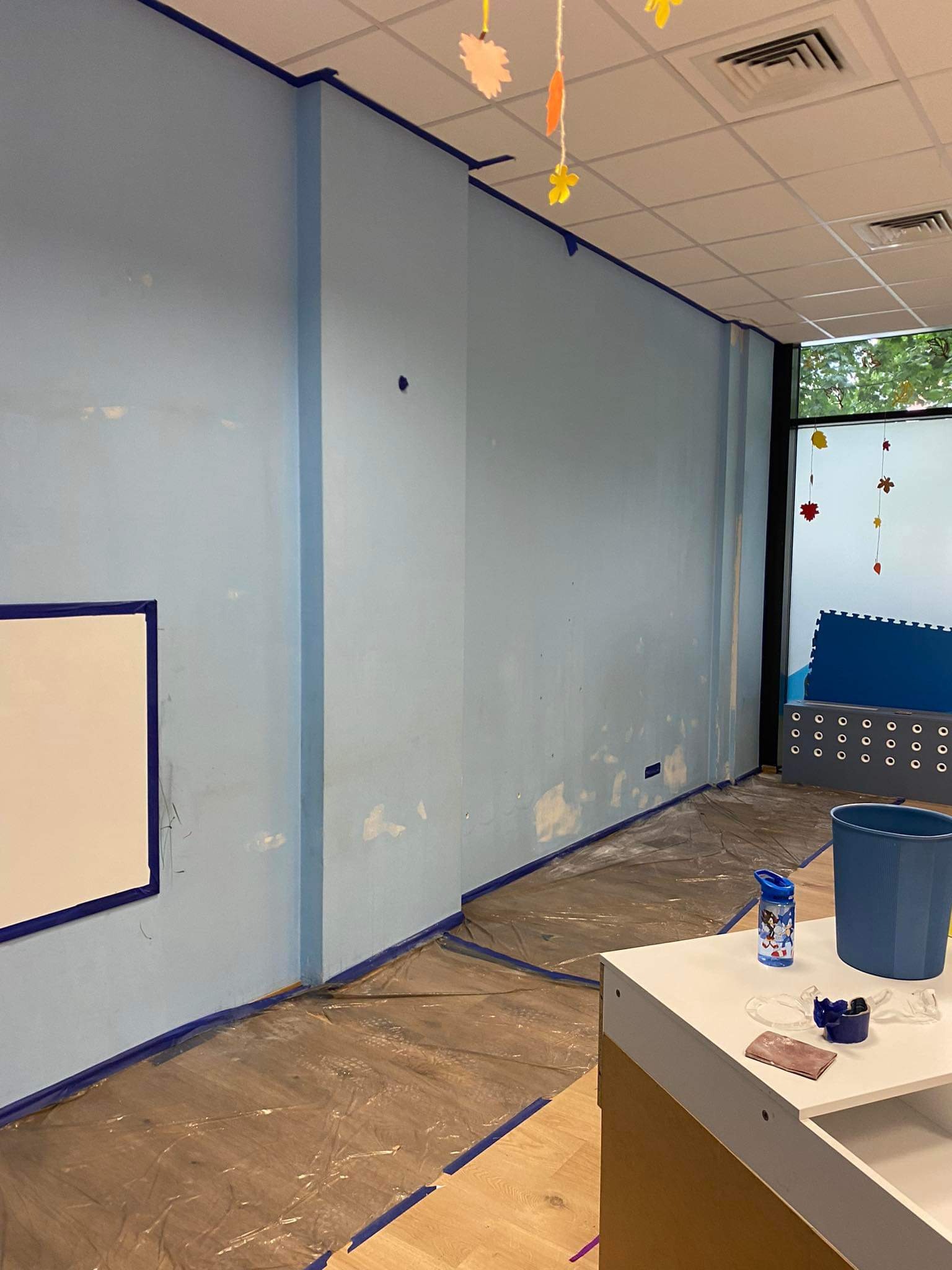 Przygotowanie ścian do malowania w pokoju dziecięcym: ściany zabezpieczone taśmą malarską, folia ochronna na podłodze, widoczne ślady po szpachlowaniu, na blacie kubek z wodą i papier ścierny.