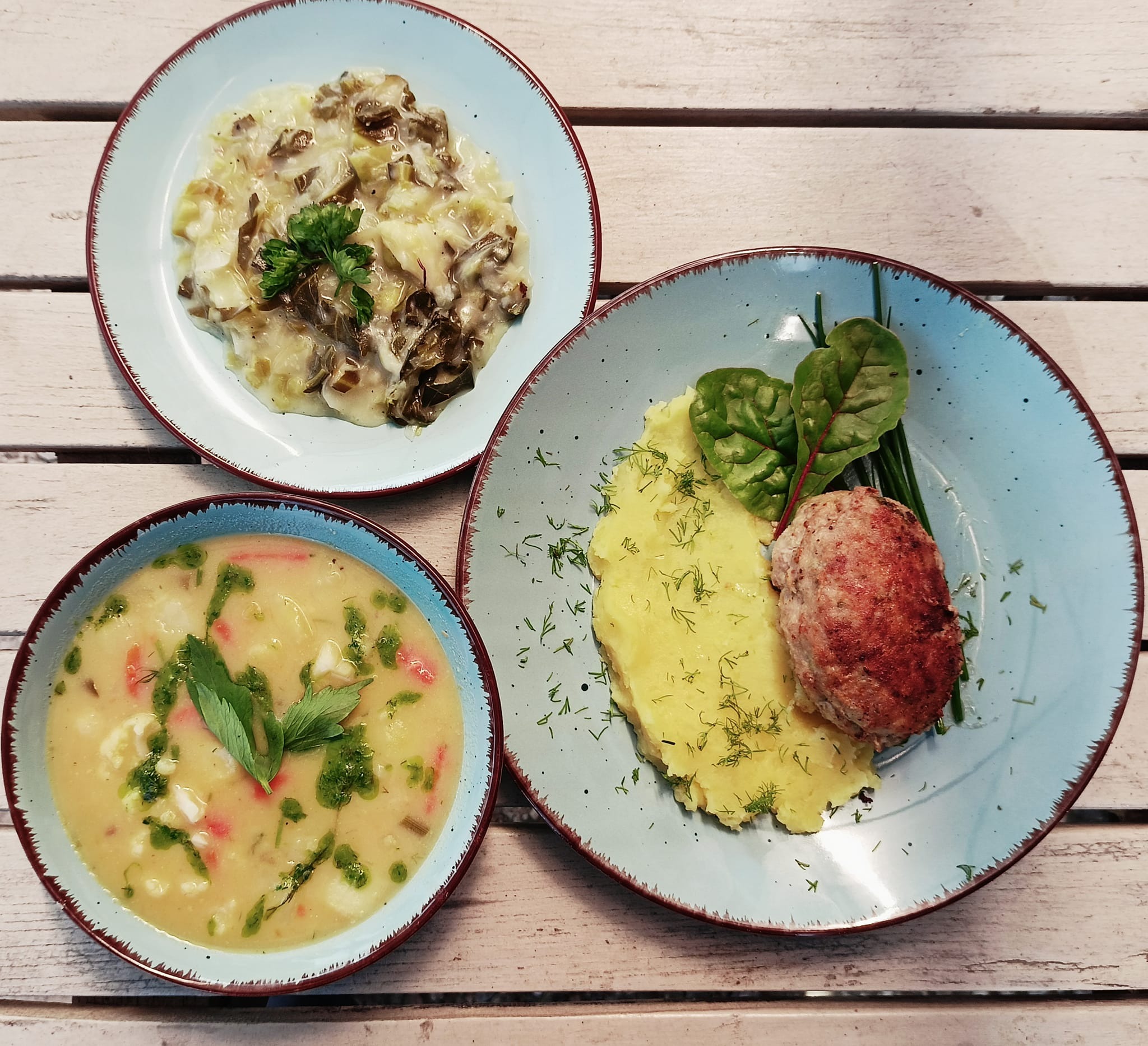 Zestaw obiadowy: zupa z zielonymi ziołami, kotlet mielony z puree ziemniaczanym posypanym koperkiem i danie z sosem grzybowym, podane na błękitnych talerzach z brązową obwódką, ułożone na białym...