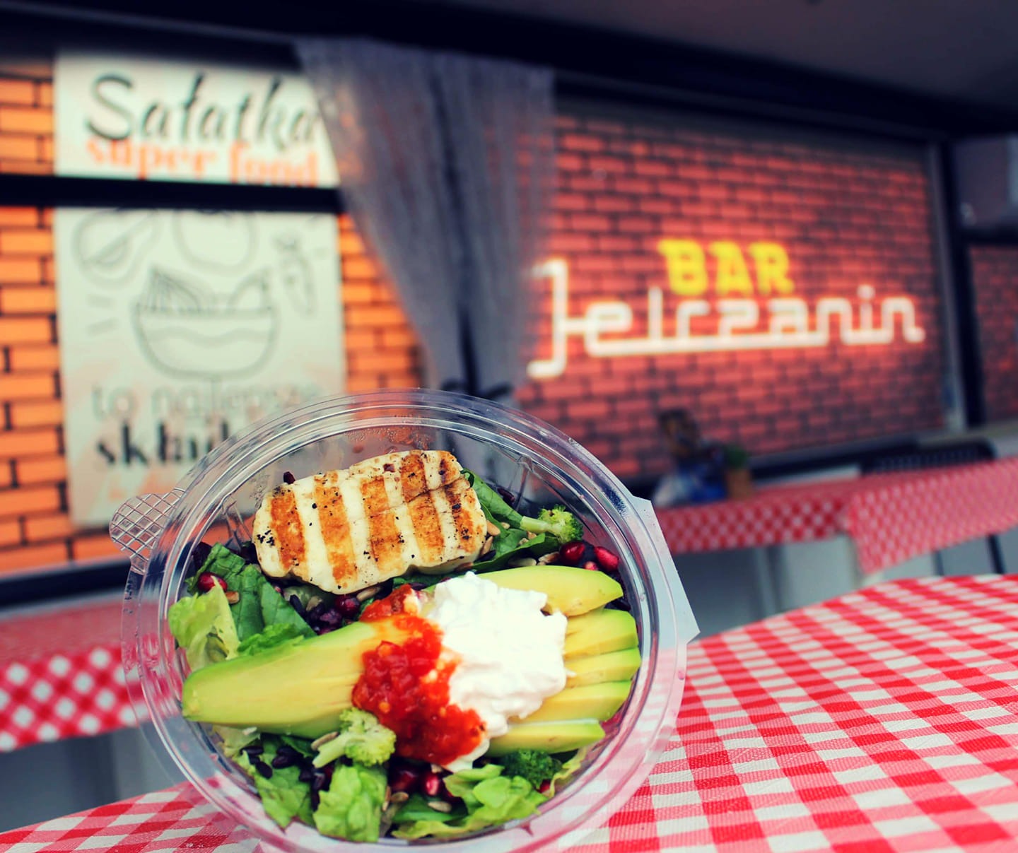 Sałatka z grillowanym serem halloumi, awokado, sosem pomidorowym i granatem w przezroczystym pojemniku na stole w czerwoną kratę, w tle neon Bar Jelczanin i plakat z napisem Sałatka Super Food.
