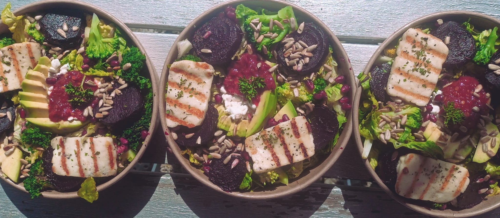 Trzy miski z sałatką: grillowany ser halloumi, buraki, awokado, brokuły, pestki słonecznika, granat i sos na jasnym, drewnianym stole.