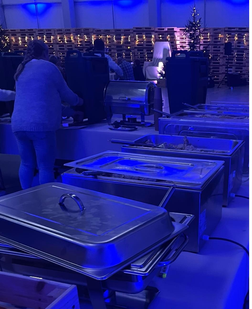 Bufet cateringowy z podgrzewaczami potraw na stole, w tle dekoracje świąteczne z palet i choinka, oświetlenie w odcieniach niebieskiego.