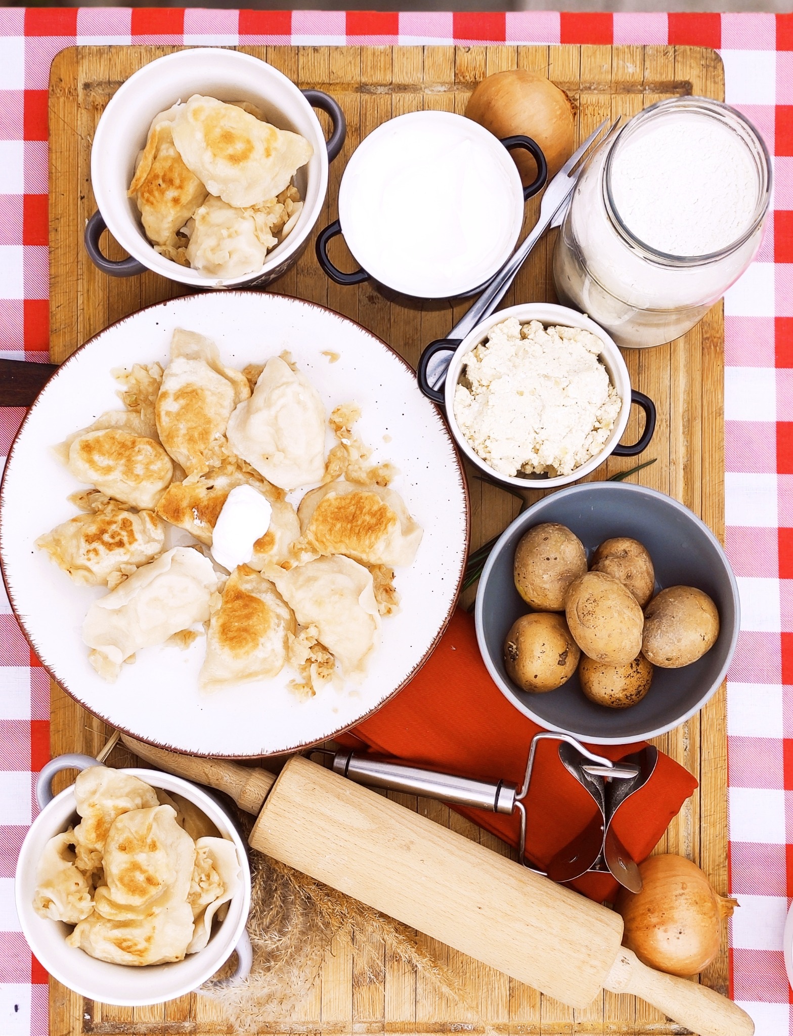 Stół nakryty w czerwoną kratę, na nim drewniana deska z pierogami, ziemniakami, serem, śmietaną, mąką, cebulą i wałkiem do ciasta, prezentacja dań w Jelcz-Laskowice.