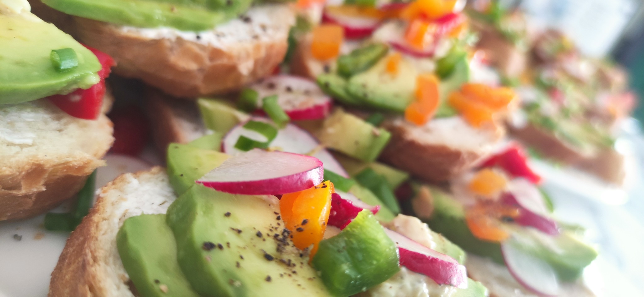 Zbliżenie na kanapki z awokado, rzodkiewką, papryką i szczypiorkiem, posypane pieprzem, na jasnym pieczywie.