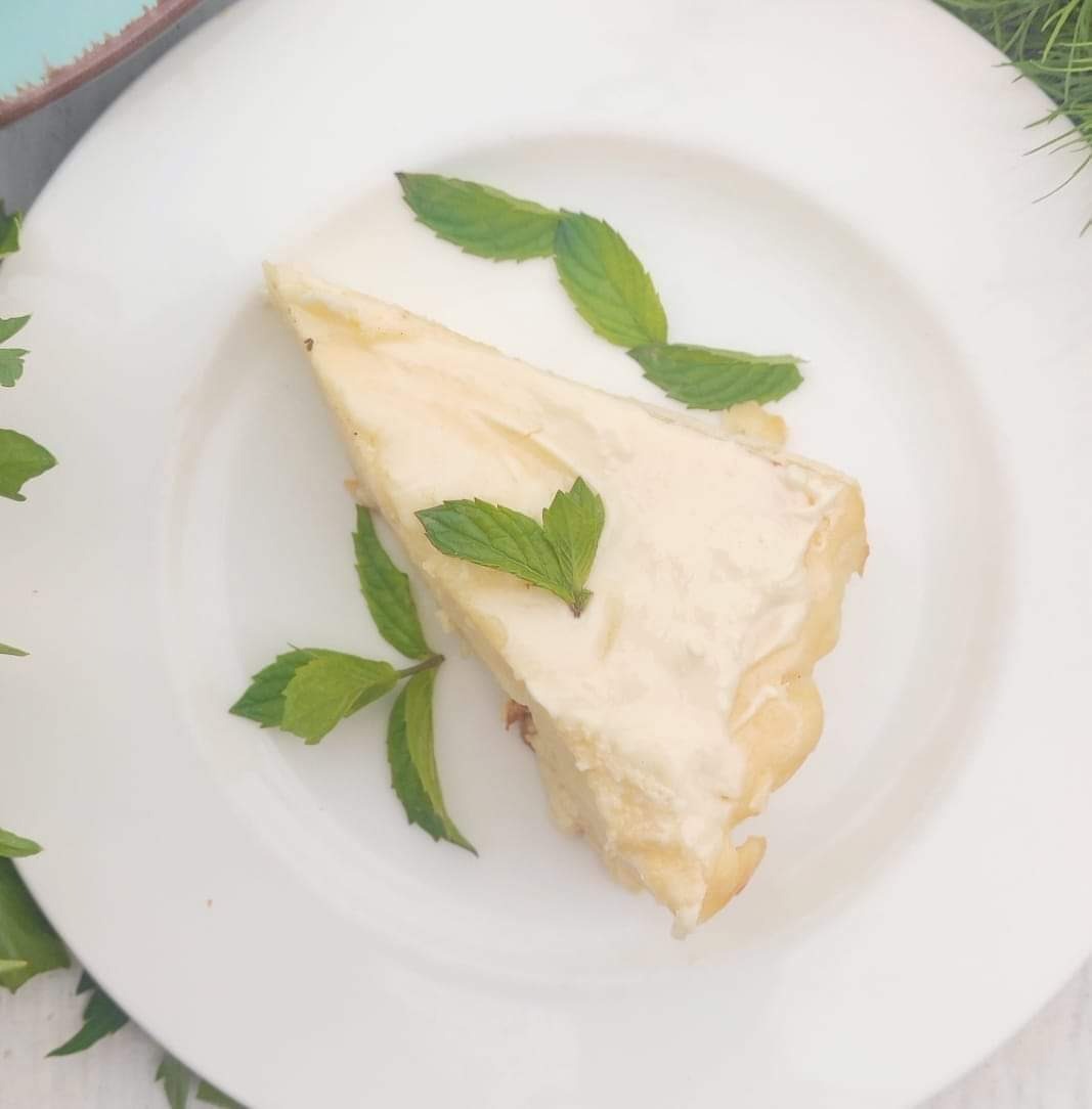 Kawałek jasnego ciasta na białym talerzu, udekorowany świeżymi listkami mięty, sfotografowany z góry w naturalnym świetle.