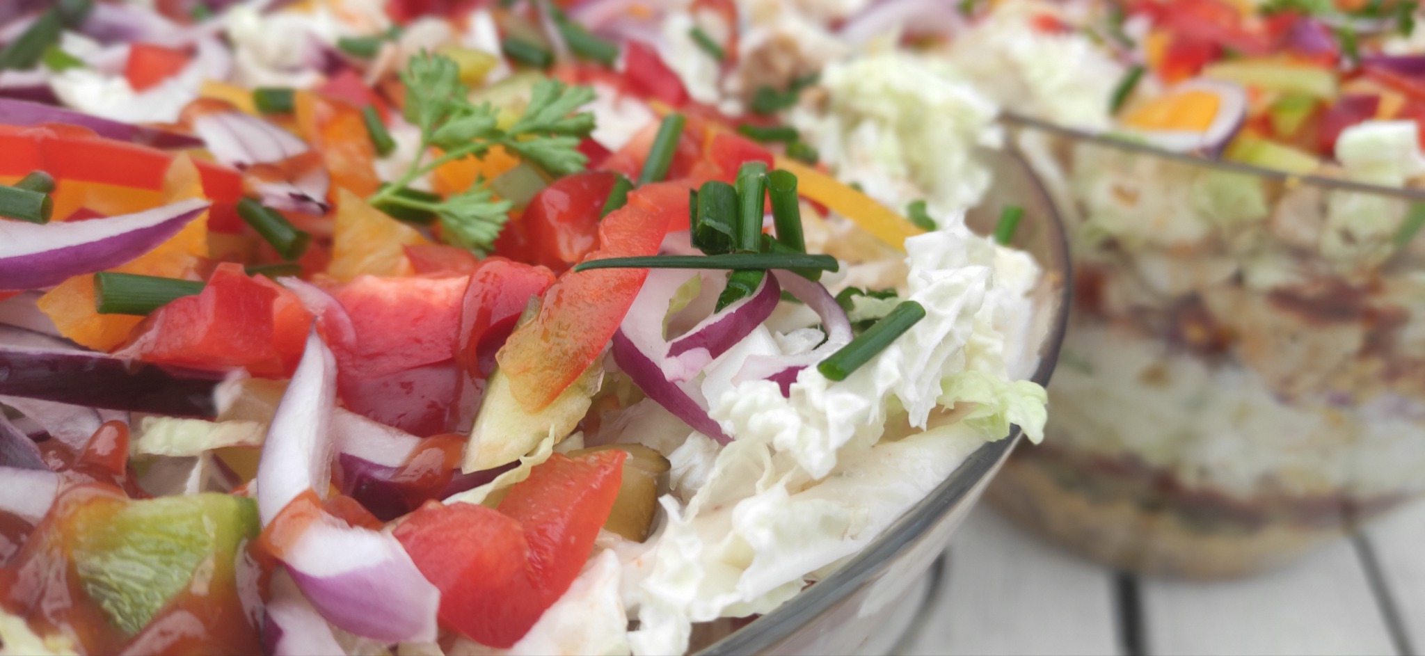 Dwie szklane misy wypełnione kolorową sałatką warzywną z czerwoną papryką, fioletową cebulą, szczypiorkiem i kapustą pekińską, ułożoną na białym drewnianym stole.