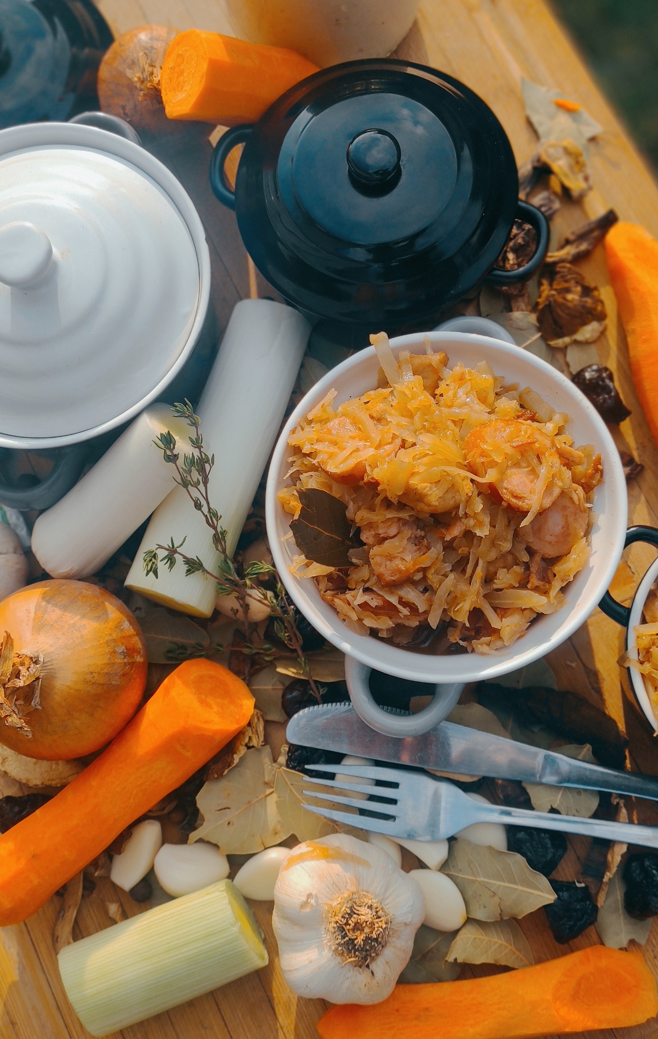 Przygotowana kapusta z kiełbasą w białej miseczce, otoczona składnikami: marchewką, porem, cebulą, czosnkiem, przyprawami i naczyniami na drewnianym stole w ciepłym świetle.