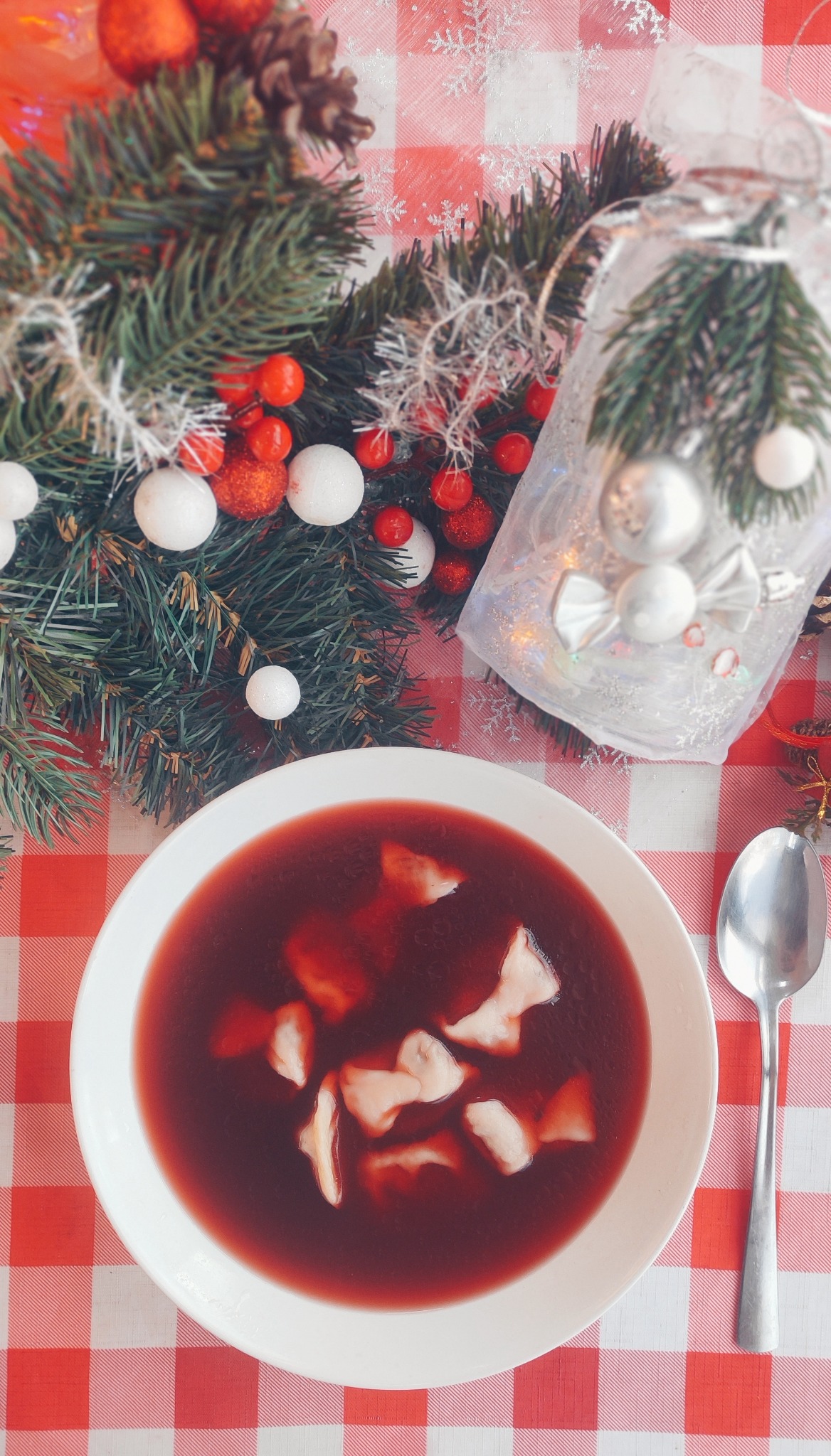 Czerwony barszcz z uszkami w białym talerzu, ozdobiony gałązkami świerku, bombkami i szyszką na czerwono-białym obrusie w kratę, obok leży srebrna łyżka.