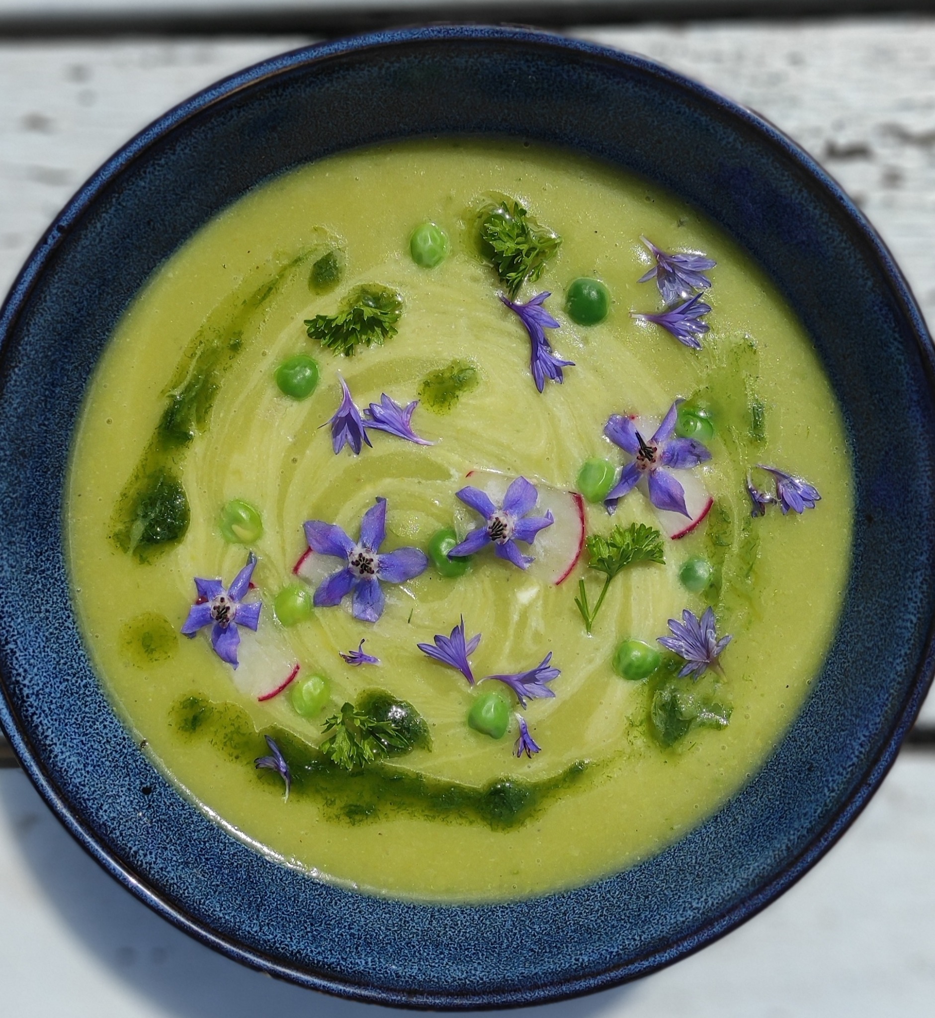 Kremowa zupa z groszku w ciemnoniebieskiej misce, udekorowana świeżymi kwiatami, groszkiem, rzodkiewką i pesto na jasnym drewnianym stole.