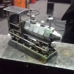 Metalowa rzeźba lokomotywy wykonana ze spawanych nakrętek i pręt&oacute;w, stojąca na metalowym stole w warsztacie.