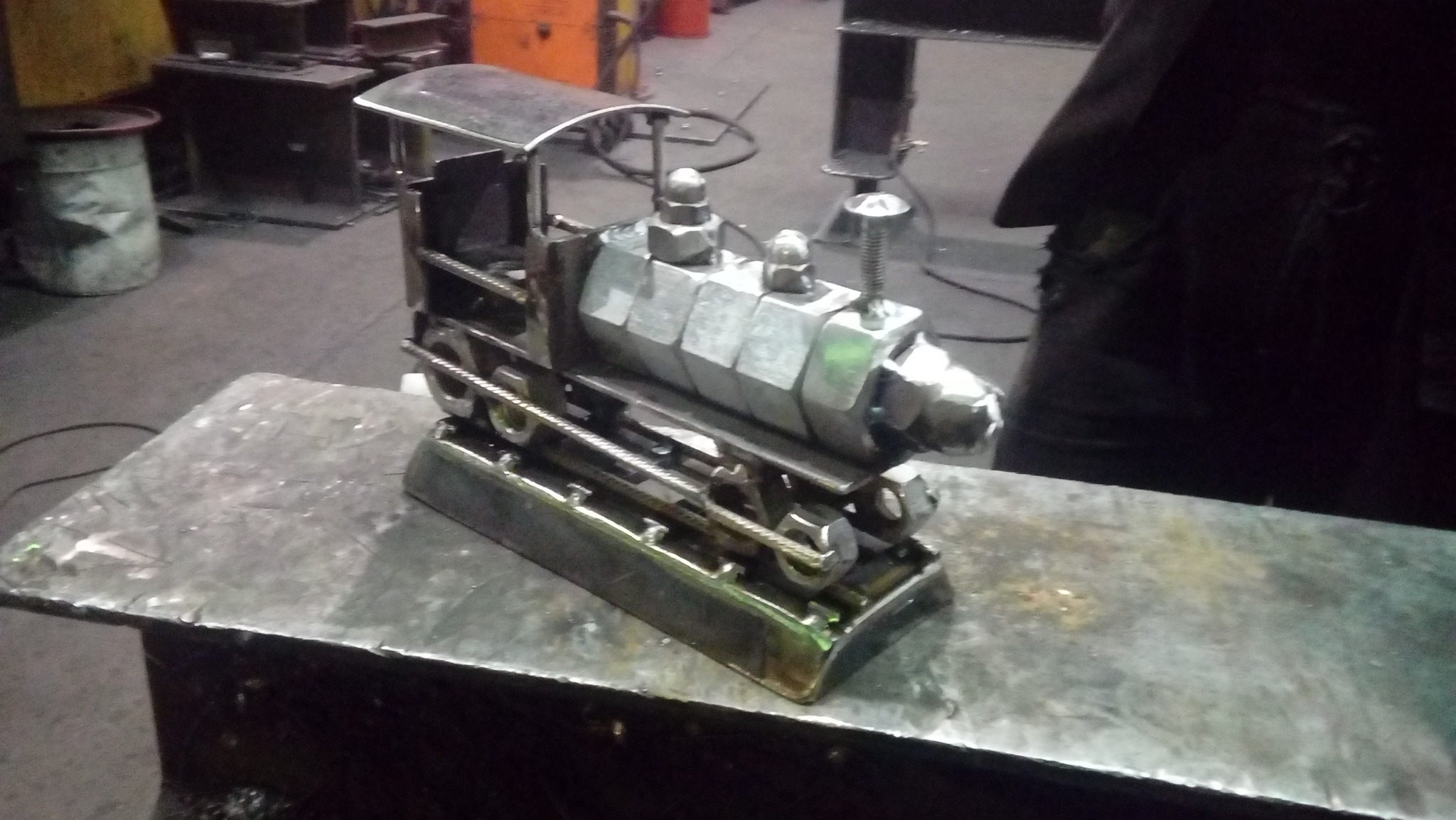 Metalowa rzeźba lokomotywy wykonana ze spawanych nakrętek i prętów, stojąca na metalowym stole w warsztacie.