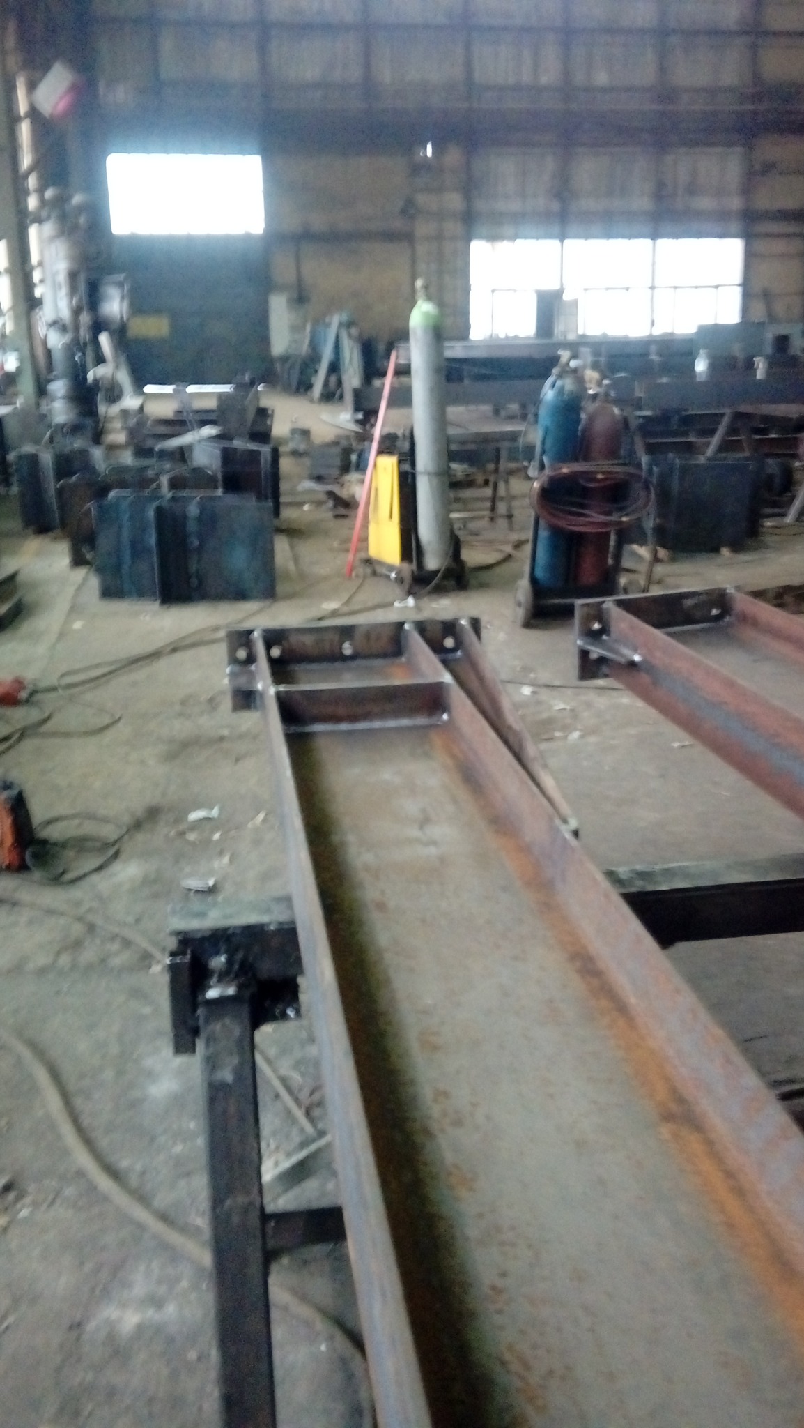 Wnętrze hali produkcyjnej z widocznymi stalowymi elementami konstrukcyjnymi na stojakach, butle gazowe do spawania oraz narzędzia w tle.