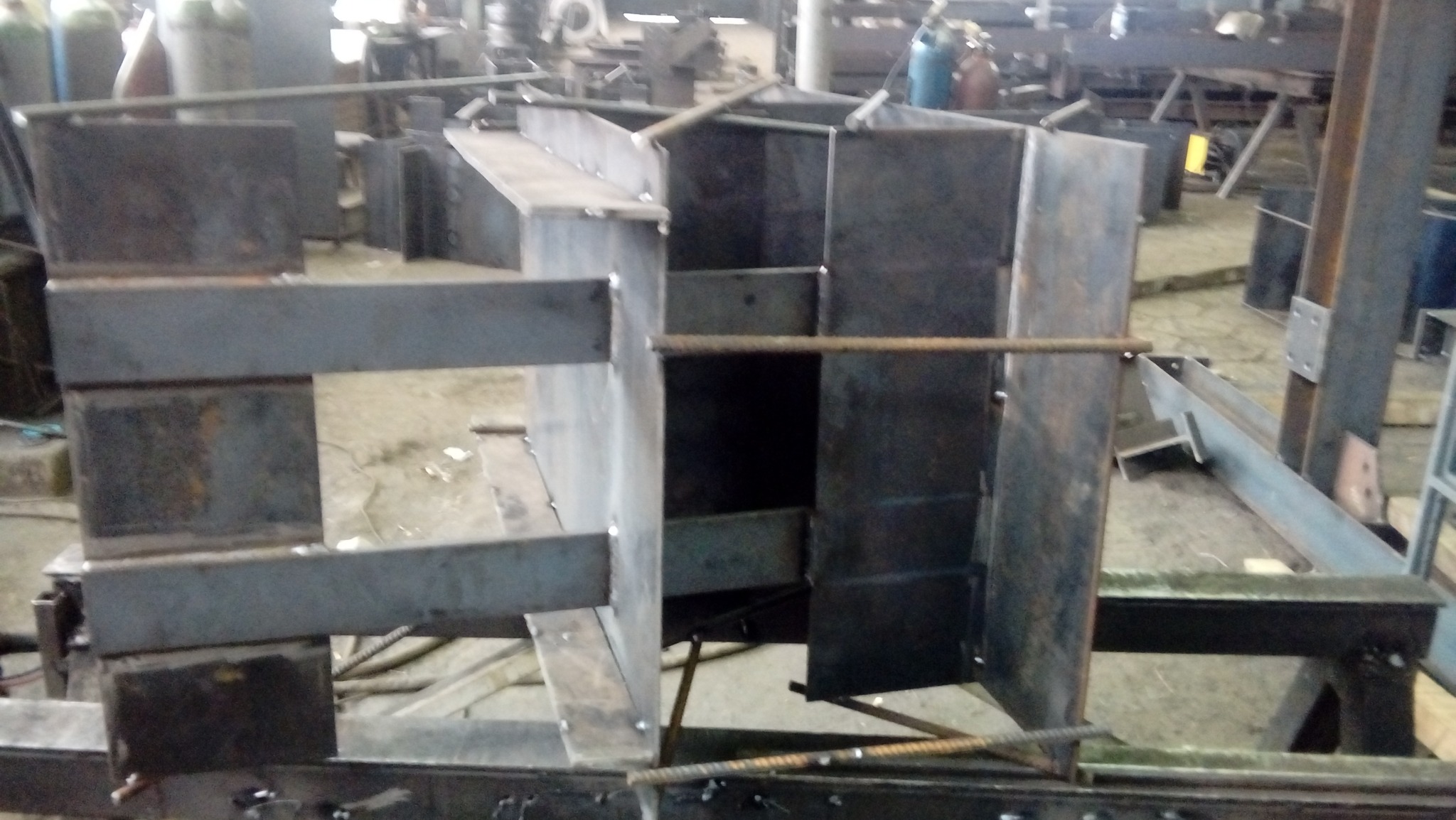 Konstrukcja stalowa w trakcie spawania, widoczne elementy z blachy i pręty zbrojeniowe, w tle warsztat.