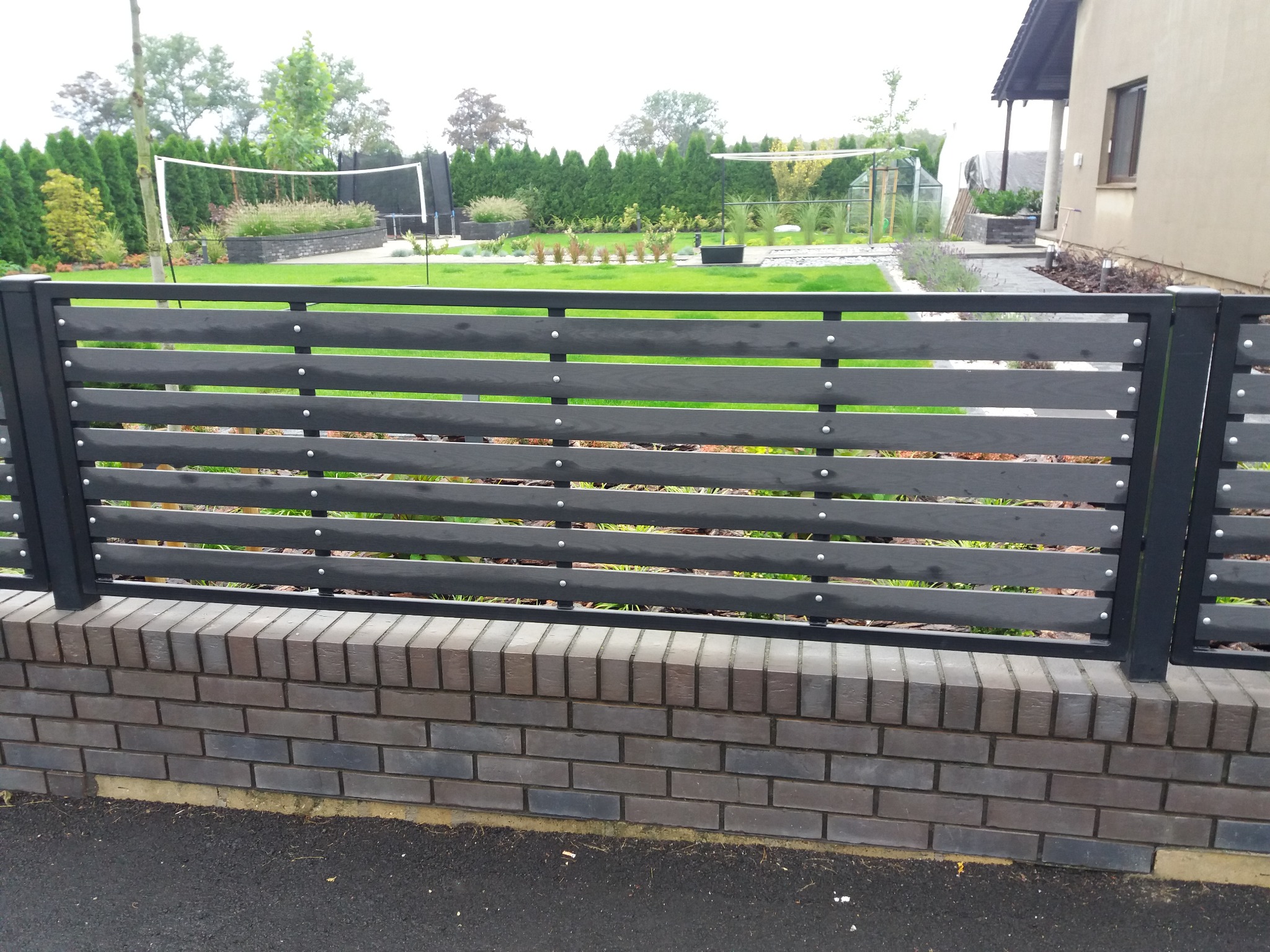 Nowoczesne ogrodzenie z poziomych, szarych desek zamocowanych na czarnych słupkach, osadzone na murku z ciemnej cegły. W tle widoczny zadbany ogród z siatką do siatkówki.